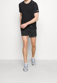 Černé tričko s krátkým rukávem od Nike s kulatým výstřihem a reflexními prvky, v kombinaci s černými kraťasy s bočním logem. Šedé sportovní boty.