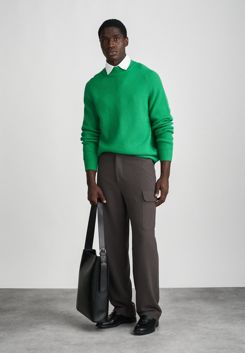 Pull vert côtelé, pantalon gris à jambes larges avec poche cargo, chaussures en cuir noires et un sac fourre-tout noir. Chemise blanche à col en dessous.