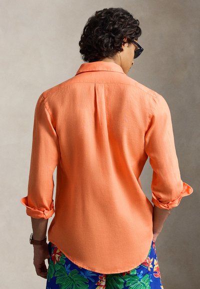 Άνδρας με σκούρα σγουρά μαλλιά που φοράει ένα πορτοκαλί πουκάμισο με τα μανίκια γυρισμένα και μπλε σορτς με λουλουδάτο σχέδιο, γυρισμένος προς τα πίσω μπροστά σε μια μονόχρωμη επιφάνεια.