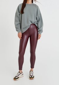Kvinna iklädd en overdimensionerad grå sweatshirt, maroon läderleggings och vita sneakers med gröna och lila accenter, stående på vit bakgrund.