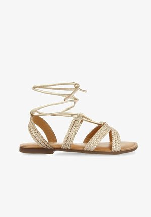 Sandalias planas trenzadas doradas con envoltura en el tobillo, que cuentan con una plantilla marrón suave y un diseño de punta abierta, enfatizando la construcción ligera.
