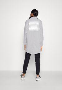 Abrigo largo de color gris claro con un diseño en la parte trasera que presenta rayas horizontales y un círculo. Combinado con pantalones jogger negros y zapatillas blancas.