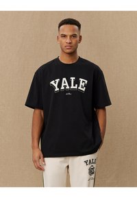 T-shirt oversized de algodão preto com "YALE" impresso em grande fonte branca. Manga curta e gola redonda. Textura suave.