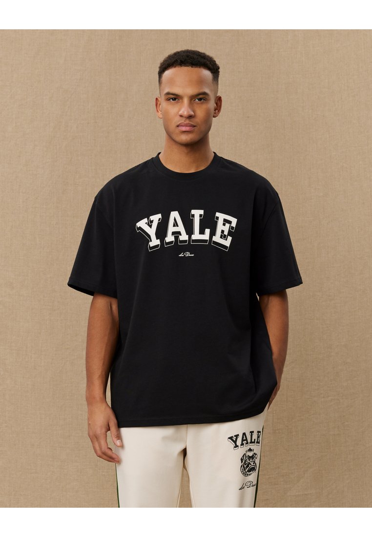 T-shirt oversized de algodão preto com "YALE" impresso em grande fonte branca. Manga curta e gola redonda. Textura suave.