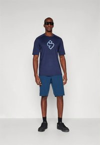 Tamsiai mėlynas sportinis marškinėlis su logotipu, derinamas su žalsvais šortais. Modelis dėvi juodus akinius nuo saulės ir tamsius sportinius batelius, stovėdamas prie balto fono.