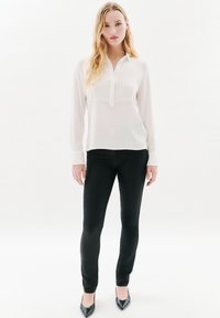 Camisa blanca de manga larga con textura acanalada vertical en el pecho, combinada con pantalones negros ajustados y tacones negros de punta. Acabado de tela suave.