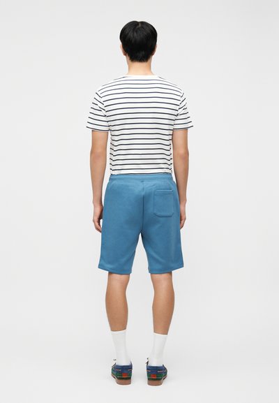 Άνδρας που στέκεται με την πλάτη στραμμένη, φοράει λευκό και ναυτικό μπλε ριγέ μπλουζάκι, μπλε σορτς με πίσω τσέπη, λευκές κάλτσες και μπλε μοκασίνια.