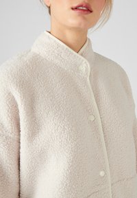 Crème-kleurige fleecejack met een gestructureerd oppervlak, staande kraag en lichtkleurige piping. Voorzien van een knoopsluiting en een zachte afwerking.