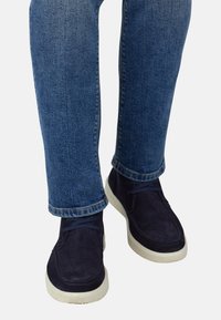 Zapatos de ante azul marino con suelas de goma blanca y cordones azul oscuro, llevados con pantalones vaqueros de mezclilla corta de color azul.
