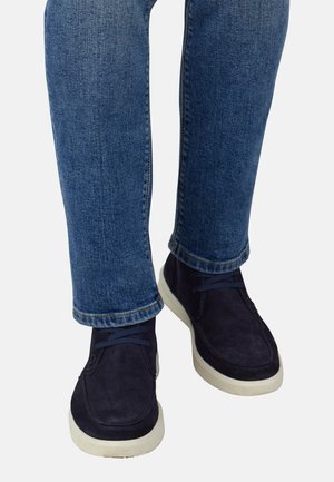 Chaussures en suède marine avec semelles en caoutchouc blanches et lacets bleu foncé, portées avec un jean en denim bleu coupé.