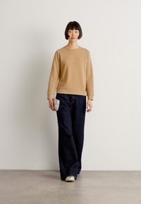 Giovane con capelli corti e scuri che indossa un felpa beige, pantaloni larghi scuri, scarpe bianche, mentre tiene una piccola pochette argentata, in piedi all'interno.
