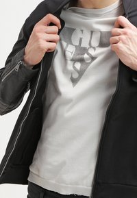 Czarna kurtka z zamkiem, nałożona na jasnoszary sweatshirt z trójkątnym logo w ciemnoszarym kolorze. Materiał wydaje się gładki i dopasowany.