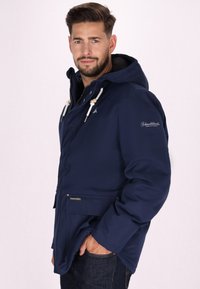 Giacca impermeabile blu navy con cappuccio, dettagli con cordoncino e tasche anteriori. Presenta una chiusura con zip e un tessuto testurizzato per calore.
