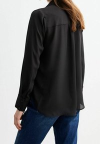 Blouse noire à manches longues avec une texture lisse, poignets boutonnés et un ourlet légèrement arrondi, associée à un jean en denim bleu.