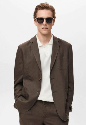 Brauner, taillierter Blazer mit zwei Knöpfen, darunter ein strukturiertes, weißes Polohemd, und schmal geschnittene Hose. Modell trägt schwarze Sonnenbrillen.