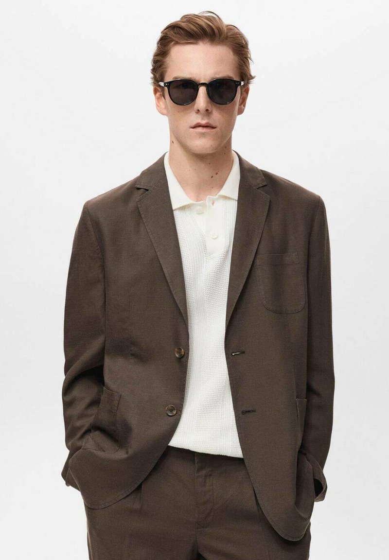 Brun skræddersyet blazer med to knapper, tekstureret hvid polo under, og slim-fit bukser. Model iført sorte solbriller.