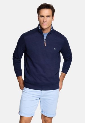 Hombre con sudadera azul marino de media cremallera y pantalones cortos azul claro remangados, de pie con las manos en los bolsillos sobre un fondo blanco.