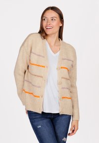 Giovane donna sorridente, che indossa un cardigan a maglia beige con strisce arancioni e viola su una camicia bianca e jeans blu strappati.