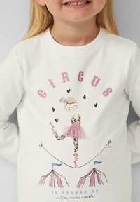 Biała bluza z napisem "CIRCUS" wykonanym z brokatowego różowego materiału. Centralna grafika przedstawia tancerkę w panterkowym wzorze z różową tiulową spódnicą, flankowaną przez namioty cyrkowe.