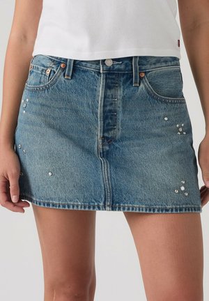 Persona che indossa una minigonna in denim blu con piccoli borchietti argento e un top bianco corto, mani rilassate ai lati.