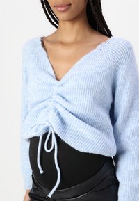 Maglione azzurro chiaro lavorato a maglia con scollatura a V profonda, con un davanti arricciato e lacci in vita. Texture morbida e dettagli di design a coste.