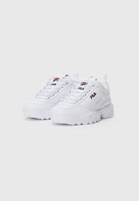 Baskets blanches FILA à semelles épaisses, logo rouge et bleu sur la languette et sur le côté, lacets blancs, présentées sur un fond léger uni.