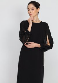 Czarna sukienka z długimi przezroczystymi rękawami i jednolitą górą. Posiada okrągły dekolt oraz ozdobny detal w postaci łańcuszka z boku.
