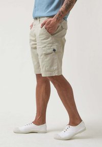 Bermudas de color beige con bolsillos laterales y detalles de botones, combinadas con zapatos de lona blancos. La tela parece ligera y casual.