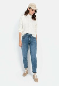 Weißer Kabelstrickpullover, hellblaue Jeans, beige Sneakers und eine beige Mütze. Die Jeans haben einen lässigen Schnitt mit aufgerollten Säumen.