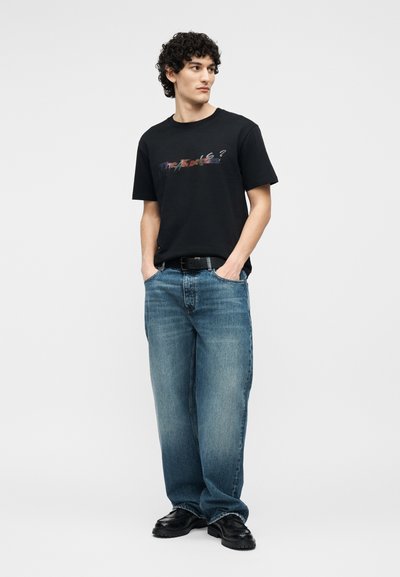 Άνδρας με σγουρά μαλλιά, που φοράει μαύρο γραφικό μπλουζάκι, φαρδιά μπλε τζιν, μαύρη ζώνη και μαύρα παπούτσια, στέκεται με τα χέρια στις τσέπες, κοιτάζοντας αριστερά.