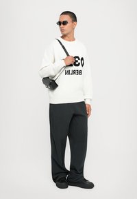 Weißer Pullover mit schwarzer "03 BERLIN" Aufschrift, kombiniert mit lockeren schwarzen Hosen und schwarzen Sneakers. Die Accessoires umfassen eine kleine schwarze Tasche und Sonnenbrille.