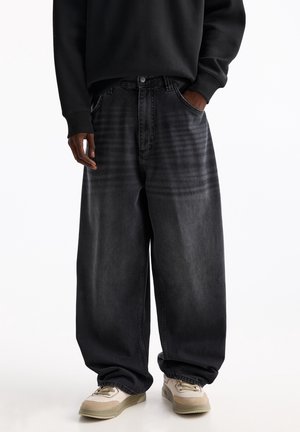 Person trägt übergroße schwarze gewaschene Jeans mit einer Hand in der Tasche und beige Sneakers auf weißem Hintergrund.
