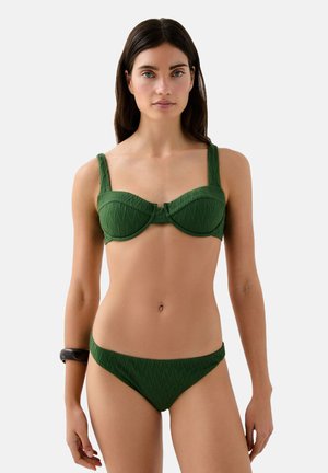 La modelo lleva un bikini verde texturizado con un top estructurado y una parte de abajo de tiro bajo, de pie frente a un fondo blanco liso.