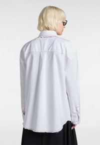 White button-up shirt with red stitching, featuring a curved hem, long sleeves, and a vent at the back with a casual fit.