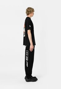 Camiseta negra con diseño gráfico en la parte trasera, combinada con pantalones negros que tienen bolsillos laterales y estampado de texto blanco. Calzado negro elegante.