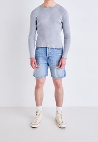 Persona de pie contra una pared lisa, vistiendo una camiseta de manga larga de canalé gris claro, pantalones cortos de mezclilla rasgados, calcetas blancas y zapatillas altas de color blanco sucio.