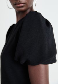 Top corto nero a maniche corte con spalle a sbuffo, realizzato in tessuto testurizzato. Primo piano che mette in risalto il design delle maniche e i dettagli morbidi e sottili.