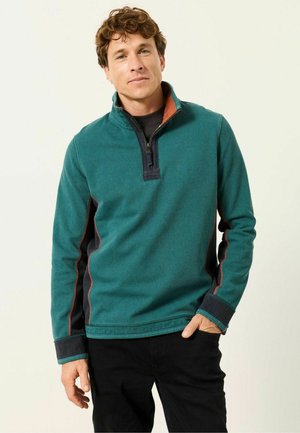Uomo con capelli castani ricci che indossa un pullover color verde petrolio con cerniera a quarto e pannelli laterali neri e arancioni e pantaloni neri, in piedi con una mano in tasca.