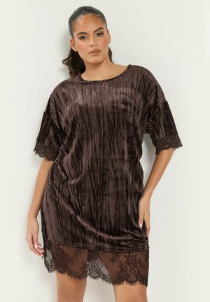 Femme portant une robe ample en velours marron foncé avec bordure en dentelle aux manches et à l'ourlet, debout devant un fond clair uni.