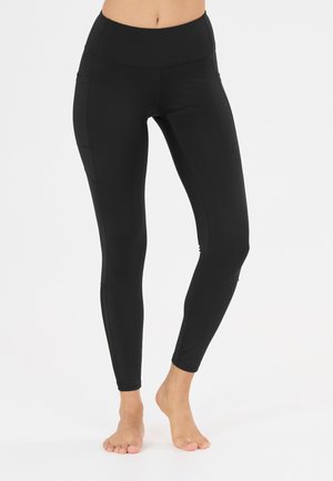Close-up van een persoon die zwarte legging met hoge taille en een zijzak draagt, blootsvoets staand op een witte achtergrond.