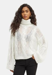 Maglione bianco a collo alto, lavorato a maglia, con motivo a trecce e trama aperta. Presenta maniche ampie e una vestibilità corta, indossato con pantaloni neri.