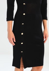 Robe en maille noire avec manches mi-longues, dotée d'une rangée de boutons dorés à l'avant et d'une fente à l'ourlet pour le détail et la facilité de mouvement.