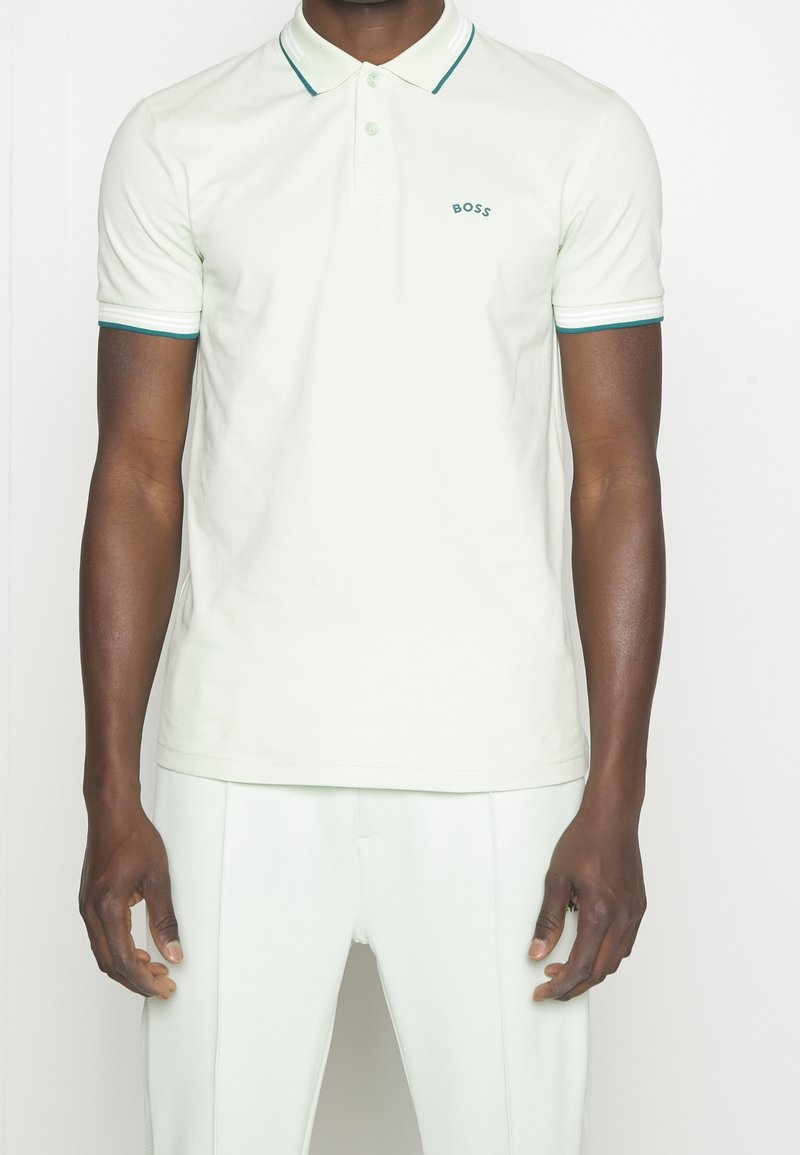 Homme portant un polo blanc avec des bordures vertes au niveau du col et des manches, et un logo vert "BOSS" sur le côté gauche de la poitrine, assorti à un pantalon blanc.