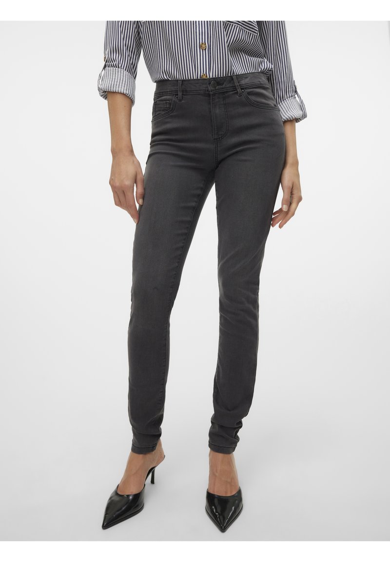 Jean gris foncé slim porté avec une chemise rayée retroussée et des talons hauts noirs pointus sur un fond uni.