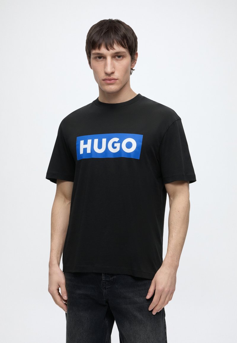 Jonge man draagt een zwart T-shirt met een blauwe rechthoek en witte "HUGO" tekst, in combinatie met een donkere spijkerbroek, tegen een effen lichte achtergrond.
