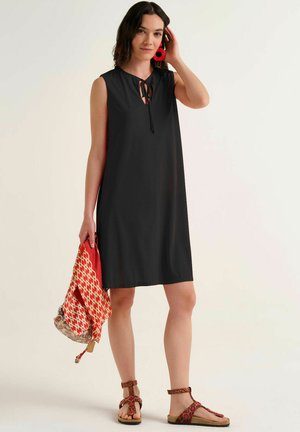 Donna che indossa un vestito nero senza maniche, orecchini rossi, che tiene una sciarpa rossa e beige con motivo e indossa sandali marroni.