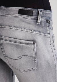 Jeans de denim gris con un corte recto, bolsillo trasero con detalles de costura y una etiqueta de logo negra en la cinturilla. Acabado texturizado.