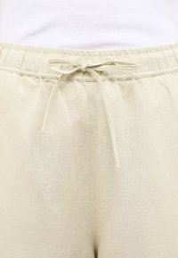 Jasno beżowe lniane spodnie z elastycznym pasem, sznurkiem do regulacji i teksturowaną powierzchnią. Materiał wydaje się miękki i przewiewny.