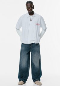 Bjela i plava prugasta košulja s kratkim rukavima, složena preko bijele majice s dugim rukavima, uparen s širokim tamnoplavi trapericama i bež cipelama.