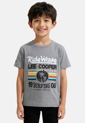 Garçon souriant aux cheveux foncés portant un t-shirt graphique gris "Ride The Wave" de Lee Cooper sur le thème du surf et un pantalon noir, debout devant un fond blanc.